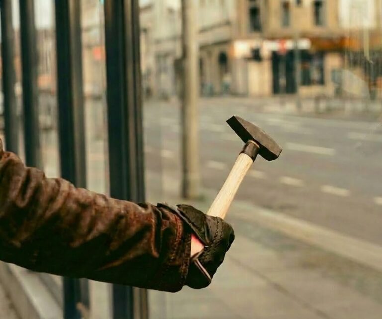 Main d’un homme tenant un marteau, sur le point de frapper la vitrine d’un magasin.